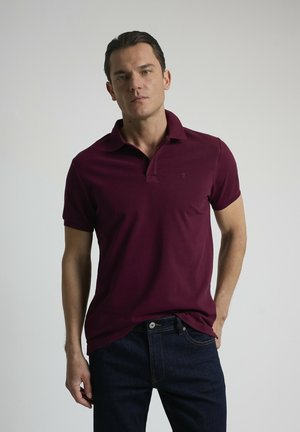 Polo burdeos de algodón suave, con manga corta, cuello clásico, que presenta un pequeño logo en el pecho, combinado con jeans oscuros.
