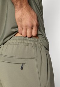 Παντελόνια αθλητικής φόρμας σε φωτεινό ελιά πράσινο με ελαστική μέση, πλαϊνή τσέπη και ραμμένα διακοσμητικά στοιχεία. Το ύφασμα φαίνεται λείο και ευέλικτο.