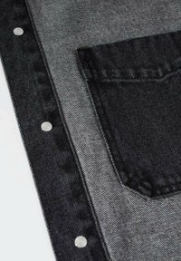 Tela de mezclilla negra con un bolsillo gris contrastante, decorado con tachuelas de metal plateado a lo largo del costado. Textura suave con un ligero brillo.