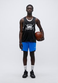 Μαύρη αμάνικη φανέλα με λευκό γραφικό, μπλε αθλητικό σορτς, μαύρα αθλητικά παπούτσια και λευκές κάλτσες. Κρατάει μια μπασκέτα.