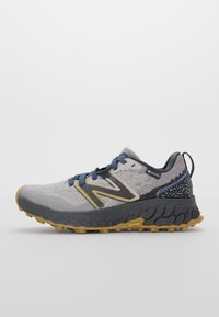 Šedá běžecká obuv s síťovaným svrškem, žlutou Vibram podrážkou, modrými tkaničkami a černým akcentem na patě. Obsahuje výrazné logo a strukturované vzory.