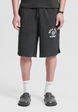 Man draagt zwarte Notre Dame-shorts, witte sokken met de tekst "RETERRNITY" en zwarte instappers, staand tegen een effen achtergrond.