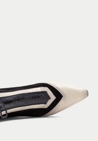 Spetsig tå-sko med en beige läderfront, svarta mockasidor och en geometrisk design. Märkbar dragkedjedetalj längs kanten.