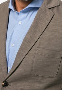 Braunes Sakko mit einer einzelnen Tasche und Knopfverschluss, kombiniert mit einem hellblauen, gemusterten Hemd mit Hemdkragen und Knöpfen.