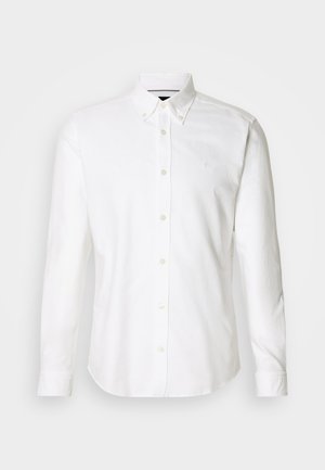 Valkoinen pitkähihainen paita, jossa on napillinen kaulus, valmistettu sileästä kankaasta, jossa on hillitty logo rinnassa ja napitus.