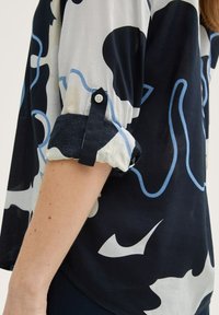 Chemise boutonnée navy et crème avec des motifs abstraits bleus, manches retroussées, tissu léger et coupe décontractée. Présente des coutures contrastées.