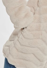 Beżowa kurtka z sztucznego futra z teksturowanym, falistym wzorem. Rękawy są puszyste, a materiał wydaje się miękki i przytulny.