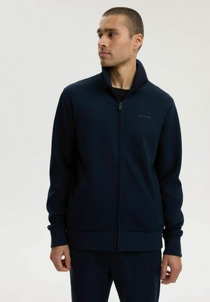 Mężczyzna w granatowej kurtce na zamek i pasujących spodniach, patrzący na bok na jednolitym jasnym tle.