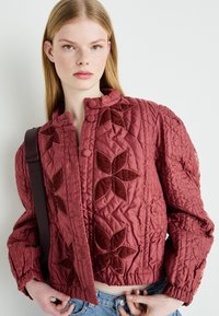 Burgundská prošívaná bunda s květinovými sametovými aplikacemi, kulatým límcem, knoflíkovým zapínáním a elastickými manžetami, doplněná o světle modré džíny.