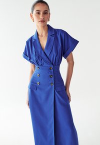 Mėlyna suknelė pagaminta iš tekstūruoto audinio, kuri turi apykaklę, trumpas rankoves, dvigubą mygtukų užsegimą ir šonines kišenes, o liemuo yra prigludęs.