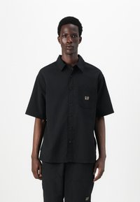 Camisa negra de botones con mangas cortas, tejido texturizado, bolsillo parche con el logo dorado "GAP", combinada con pantalones negros a juego.