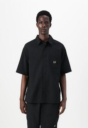 Black button-up shirt with short sleeves, textured fabric, patch pocket with gold "GAP" logo, worn with matching black pants.