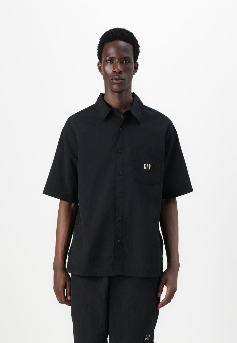 Camisa negra de botones con mangas cortas, tejido texturizado, bolsillo parche con el logo dorado "GAP", combinada con pantalones negros a juego.