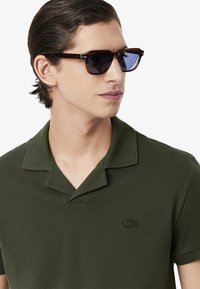 Mörkgrön polo skjorta med en strukturerad tyg, utrustad med krage och en broderad logotyp på bröstet. Bärs med mörka solglasögon.