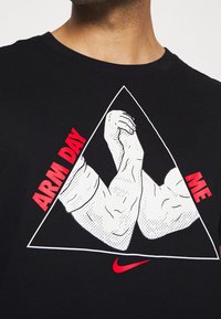 Camiseta negra con un gráfico triangular que presenta brazos musculosos ilustrados. Texto "ARM DAY ME" en rojo, logotipo de Nike en rojo en la parte inferior.