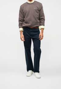 Homme portant un sweat-shirt marron Tommy Jeans, manches de chemise jaunes retroussées, pantalon bleu foncé, ceinture marron et baskets blanches, debout devant un fond blanc.