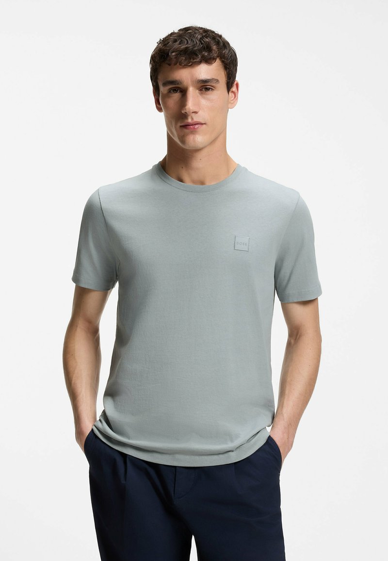 Jeune homme aux cheveux courts et bouclés, portant un t-shirt gris clair à manches courtes et un pantalon foncé, debout les mains dans les poches.
