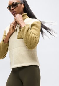 Φλις πουλόβερ σε μπεζ και ελιά, με γιακά με φερμουάρ, μπροστινή τσέπη και υφασμάτινη υφή για ζεστασιά. Φοράγεται με κολάν σε απόχρωση ελιάς.