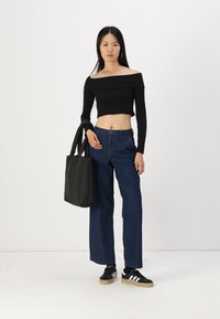 Černý žebrovaný crop top s odhalenými rameny, modré široké džíny, černá kabelka a černé tenisky s bílými pruhy, na jednoduše bílém pozadí.