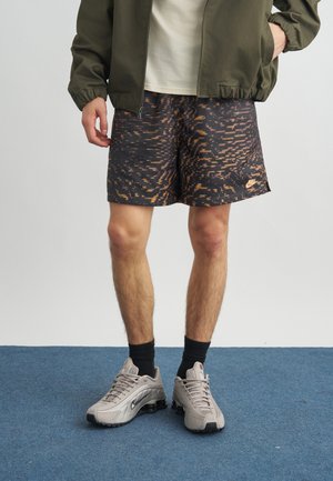 Homme portant un short Nike noir et orange à motifs, une veste olive, des baskets beige et des chaussettes noires, debout sur un tapis bleu.