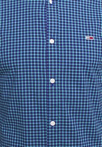 Camicia a maniche corte a quadretti blu con una combinazione di quadrati blu scuro e blu chiaro. Presenta bottoni bianchi e un piccolo logo sul petto.