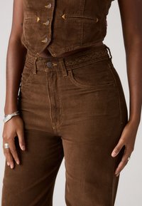 Bruine corduroy cropped vest met gouden knopen en twee zakken, gecombineerd met een hoog-waisted bijpassende corduroy broek met een gevlochten tailleband en zichtbare naden.