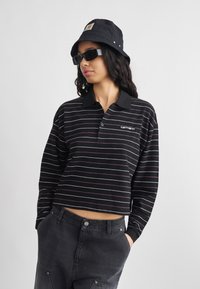 Νεαρή γυναίκα με μαύρο καπέλο τύπου bucket, μαύρα γυαλιά ηλίου, μακρυμάνικο cropped τοπ με ασπρόμαυρες ρίγες και σκούρο τζιν, με τα χέρια στις τσέπες.