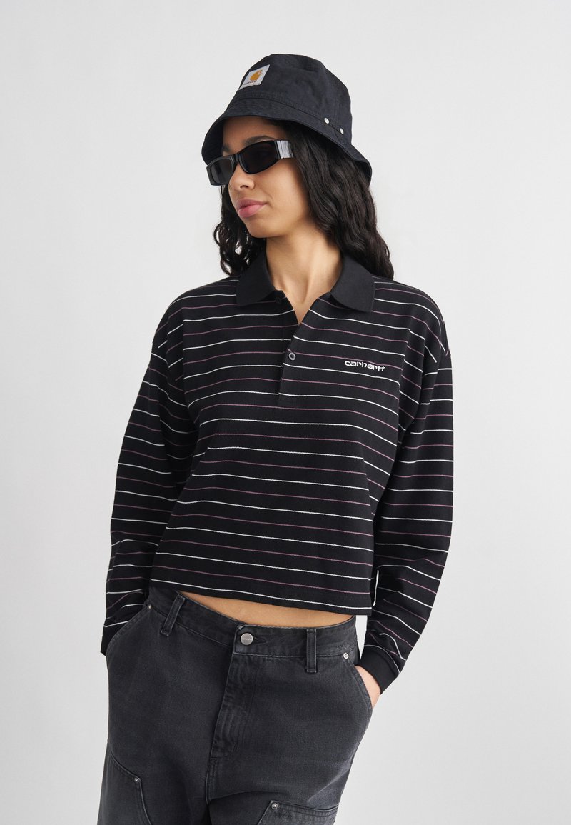 Νεαρή γυναίκα με μαύρο καπέλο τύπου bucket, μαύρα γυαλιά ηλίου, μακρυμάνικο cropped τοπ με ασπρόμαυρες ρίγες και σκούρο τζιν, με τα χέρια στις τσέπες.