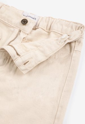 Close-up van de tailleband van beige broek met een gedeeltelijk open rits, knoop en elastische achterkant met een zichtbaar kledinglabel.