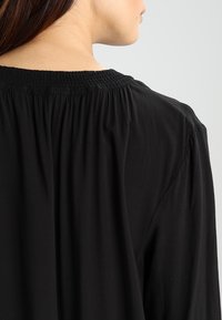 Svart blus med rundad halsringning, elastiskt smockdetalj bak, och flödig tyg med en mjuk textur.