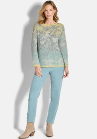 Vrouw met schouderlang blond haar, gekleed in een geel-blauwe bloementrui, lichtblauwe broek en beige enkellaarsjes, staand tegen een witte achtergrond.