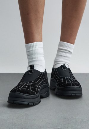 Schwarze Slipper aus Wildleder mit weißen Ziernähten. Dicke schwarze Sohlen mit Profilmuster. Getragen mit weißen, gerippten Knöchelsocken.