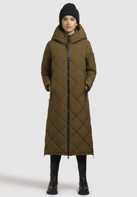 Lang, olivengrøn pufferjakke med diamantquiltet design, høj krave, fuld lynlås, sidelommer og hætte, parret med sorte støvler.