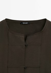 Haut marron foncé avec un col rond, une courte patte de boutonnage avec deux boutons noirs, et un tissu subtilement texturé. Étiquette visible au col.