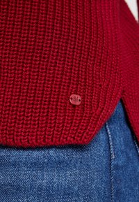 Rode gebreide trui met een geribbelde textuur, voorzien van een kleine bruine logoknop aan de zoom, gecombineerd met denim stof in blauwtinten.