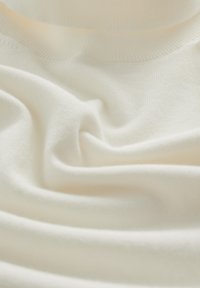 Weicher, cremefarbener Stoff mit glatter Textur, der sanfte Falten und einen gerippten Halsausschnitt für zusätzlichen Detailreichtum aufweist.