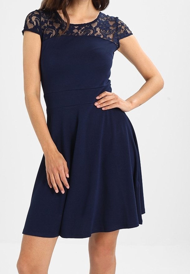 Femme portant une robe bleu marine longueur genou avec des manches courtes en dentelle et corsage, posant avec une main sur la hanche et l'autre reposant sur la jupe.