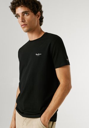 Mladý muž s kučeravými vlasmi v čiernom tričku Pepe Jeans a béžových nohaviciach, stojaci s jednou rukou v vrecku na jednolennom pozadí.
