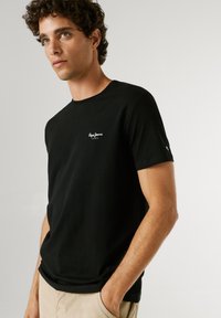 Junger Mann mit lockigem Haar, der ein schwarzes Pepe Jeans T-Shirt und beige Hose trägt, stehend mit einer Hand in der Tasche vor einfachem Hintergrund.