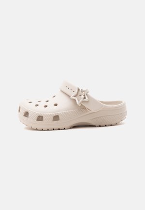 Zoccolo beige senza lacci con fori di ventilazione, suola texturizzata, cinturino regolabile e ciondolo a forma di stella su uno sfondo chiaro e uniforme.