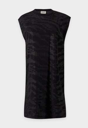 Femme portant une robe noire sans manches avec une ceinture cloutée, des bottines noires, des boucles d'oreilles argentées et tenant un sac à main noir sur un fond uni.
