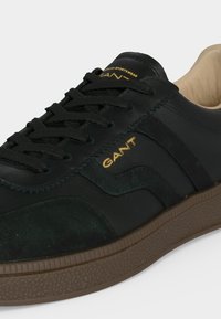 Baskets en cuir noir et daim avec semelle marron texturée, lacets noirs et logo doré GANT sur le côté et la languette.