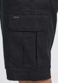 Shorts cargo noirs en tissu léger, présentant une grande poche latérale, des coutures visibles et une petite étiquette de marque à l'avant.