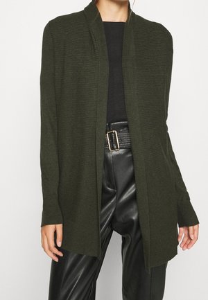 Person wears dark green open-front cardigan over black top and black high-waisted leather pants with wide belt and gold buckle.