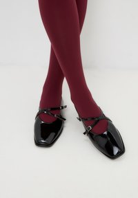 Zwarte lakleren Mary Jane-schoenen met vierkante neuzen, voorzien van twee dunne kruisbanden met gespen. Draagt met bordeauxkleurige ondoorzichtige tights.