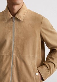 Bruine suède jasje met een ritssluiting aan de voorkant, puntige kraag, zijzakken en een zachte textuur. Heeft een minimalistisch ontwerp en subtiele stikdetails.