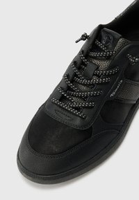 Scarpa sportiva nera con tomaia in pelle texturizzata, inserti in mesh, lacci riflettenti e suola in gomma con design a punta arrotondata.