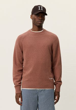 Maglione marrone lavorato a trama con collo rotondo, orlo a costine e maniche raglan; abbinato a pantaloni a righe e un berretto scuro con scritte bianche.