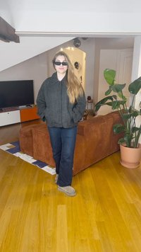 Giacca in pied de poule bianco e nero oversize, jeans blu, pantofole grigie; in piedi in una stanza con un divano in ciniglia marrone e piante.