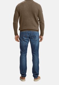 Uomo che indossa un maglione marrone, jeans blu e scarpe marroni, in piedi rivolto di spalle contro uno sfondo bianco semplice.